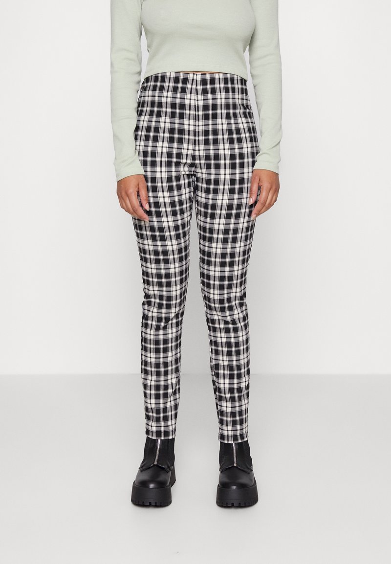 Personne portant un pantalon à carreaux noir et blanc, un crop top à manches longues gris clair et des bottes noires épaisses, debout devant un fond clair uni.