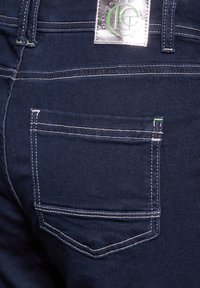 Jeans in denim blu scuro con una tasca posteriore caratterizzata da cuciture bianche e un accento verde. Un'etichetta con logo argento è attaccata alla vita.