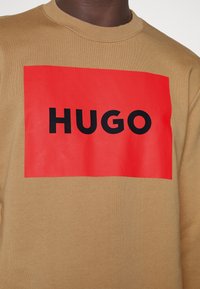 Sudadera beige con un gran estampado rectangular rojo que presenta el texto negro y audaz "HUGO" en el pecho.