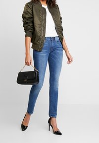 Chaqueta bomber verde oliva, camiseta blanca, jeans ajustados azules, bolso de cuero negro con cadena dorada y tacones negros de punta.