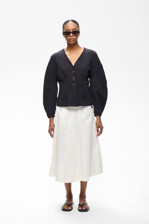 Femme portant une veste noire boutonnée avec un col en V, une jupe midi plissée blanche, des sandales noires et des lunettes de soleil foncées, debout sur un fond blanc.