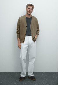 Cardigan lavorato a maglia marrone sopra una t-shirt grigia, abbinato a pantaloni bianchi e scarpe scure. Lo sfondo presenta un'ambientazione minimalista.