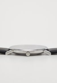 Sølv rustfritt stålklokke med en glatt rund urskive, svart lærband og en fremtredende krone på siden. Enkel, minimal design.