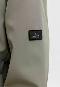 Kaki látka s čiernou obdĺžnikovou nášivkou s logom Jack & Jones Premium vysokokvalitne prišitou na rukáve, zobrazujúca textúrované detaily textílie.