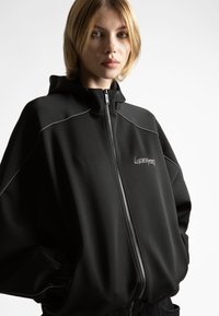 Fiatalember rövid szőke hajjal, fekete, fehér díszítésű cipzáras kapucnis pulóverben, hódolattal kezével a zsebében.