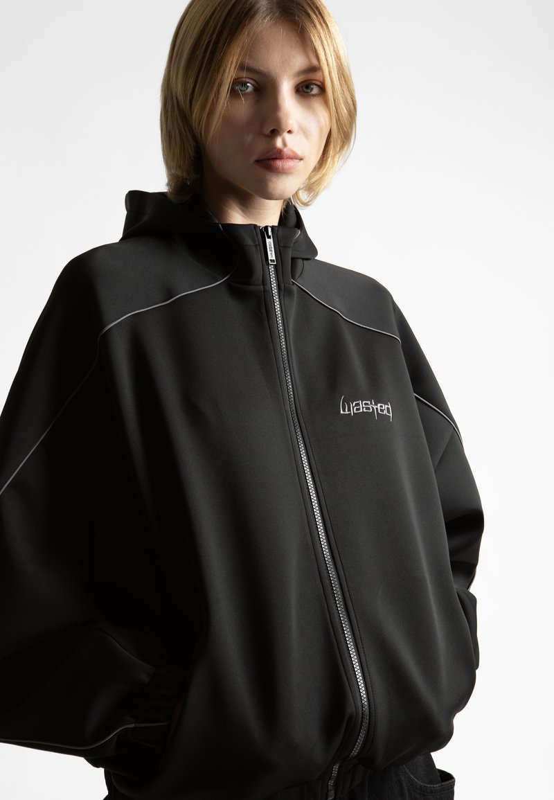 Junge Person mit kurzen blonden Haaren, die eine schwarze Kapuzenjacke mit weißer Paspel und einem gestickten Logo auf der Brust trägt, Hände in den Taschen.