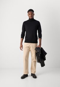 Suéter negro de cuello alto, pantalones color crema y botas de cuero negras. Sosteniendo una chaqueta de cuero negra. Texturas suaves, diseño ajustado.
