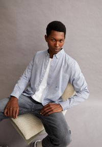 Lichtblauw overhemd met knopen en een klein logo, gedragen over een wit t-shirt, gecombineerd met grijze jeans. Model zit op een textuurstoel.