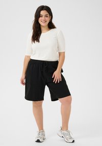 Haut blanc à rayures, à manches trois-quarts, associé à un short noir à taille élastique avec poches. Le modèle porte des baskets gris clair.