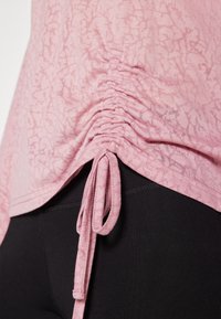 Pink top with a textured pattern, featuring gathered sides and adjustable ties. Black leggings are partially visible beneath.