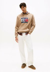 Giovane uomo che indossa un hoodie beige di Tommy Jeans, una camicia a quadri sotto, pantaloni bianchi e sneakers marroni, in piedi con una mano in tasca.
