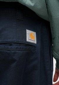 Granatowe spodnie Carhartt z wszywaną metką z pomarańczowym logo na szarym tle. Materiał to wytrzymały denim o gładkiej fakturze.