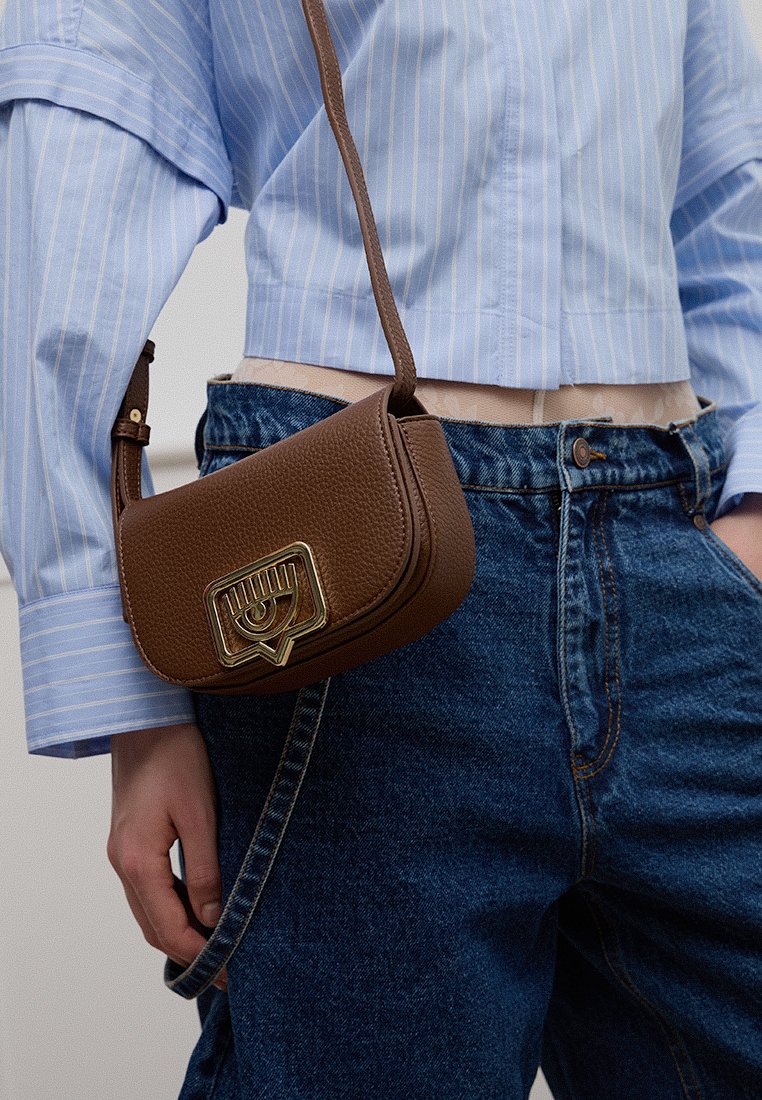 Bolso crossbody de cuero marrón con un acabado texturizado, herrajes dorados y un acento en forma de ojo único. Llevado con una camisa a rayas azul y vaqueros.