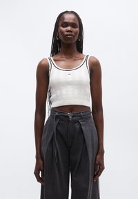 Bílý žebrovaný krátký top s námořnicky modrým lemováním; zdobený texturovaným vzorem a logem. Kombinováno s tmavě šedými kalhotami s vysokým pasem a sklady.