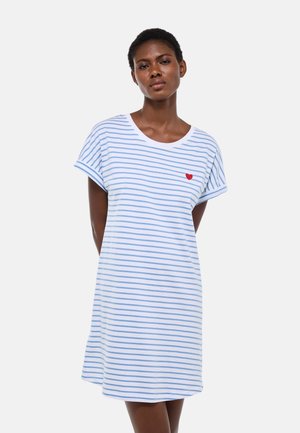 Femme portant une robe blanche à rayures horizontales bleu clair et un petit cœur rouge sur le côté gauche de la poitrine, debout avec les mains derrière le dos.