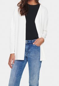 Kvinna som bär en lång vit öppen kofta över en svart topp och blåjeans, med ena handen i fickan och den andra avslappnad vid sin sida.