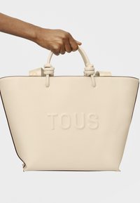 Beżowa torba na ramię wykonana z teksturowego materiału, z wytłoczonym logo "TOUS" i podwójnymi węzłowymi uchwytami, z kontrastowym brązowym wykończeniem.