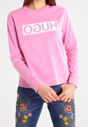 Felpa rosa realizzata in morbido materiale con un grande logo bianco "HUGO" sulla parte anteriore; indossata con jeans blu decorati con ricami floreali.