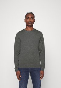 Maglione lavorato a maglia verde scuro con scollatura rotonda e maniche lunghe, caratterizzato da un tessuto strutturato e orlo a coste. Indossato con jeans blu.