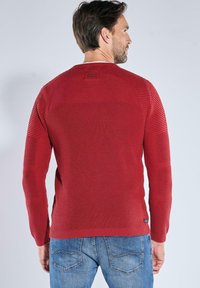 Roter gerippter Strickpullover mit kontrastierenden strukturierten Einsätzen und rundem Halsausschnitt, kombiniert mit blauen Jeans. Verfügt über ein Logo-Tag nahe dem Saum.