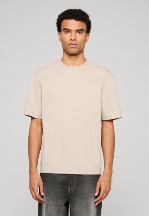 Man with short curly hair wearing plain beige short-sleeve t-shirt and faded black jeans, standing against a plain light gray background.