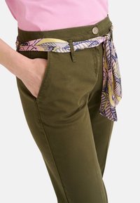 Olivgrüne maßgeschneiderte Hose mit Seitentaschen, ausgestattet mit einem gemusterten pink-blauen Gürtel, der in der Taille gebunden ist. Glatte Stofftextur.