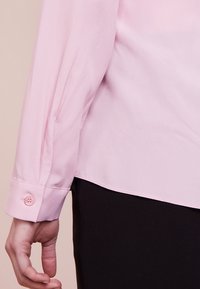 Persoon in een lichtroze shirt met lange mouwen en knoopmanchetten, en zwarte broek, met een ontspannen hand langs hun zij tegen een neutrale achtergrond.