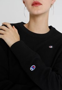 Felpa nera con polsini e scollo a costine, con piccolo logo ricamato in rosso e blu sul petto e sulla manica sinistra.