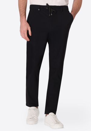 Uomo che indossa pantaloni casual neri con coulisse e sneakers bianche, in piedi con le mani vicino alle tasche su sfondo semplice.