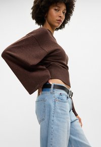 Suéter de malha castanho brilhante com mangas largas, combinado com jeans de cintura alta em azul claro e um cinto preto, destacando um look casual.