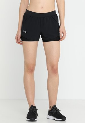 Dolna część ciała osoby ubranej w czarne, warstwowe krótkie spodenki sportowe oraz czarne buty do biegania, stojącej na jednolitym białym tle.