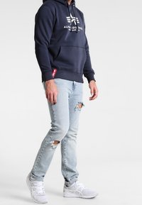 Felpa blu navy con logo frontale, cappuccio con cordino e tasca a marsupio, abbinata a jeans chiari strappati e sneakers bianche.