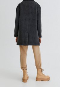 Czarny i szary płaszcz w kratę z długimi rękawami, beżowe spodnie zwężane oraz beżowe sznurowane buty z masywną podeszwą i detalem na zamku.