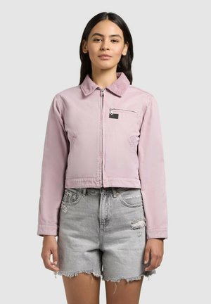 Nő világos rózsaszín, cipzáras, galléros dzsekiben és szürke, kopott farmer rövidnadrágban, egyszínű háttér előtt áll.