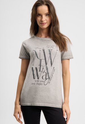 Frau mit langen braunen Haaren, die ein hellgraues T-Shirt mit dem Aufdruck "new way write your next chapter" und schwarze Hosen trägt.