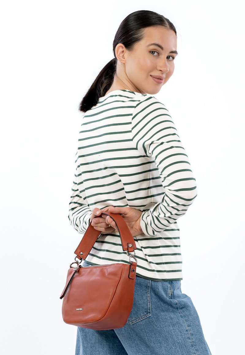 Brūna ādas soma ar īsu rokturi, apaļu dizainu un smalkām šuvju detaļām, ko tur cilvēks, kurš valkā svītrainu garām piedurknēm jaku.