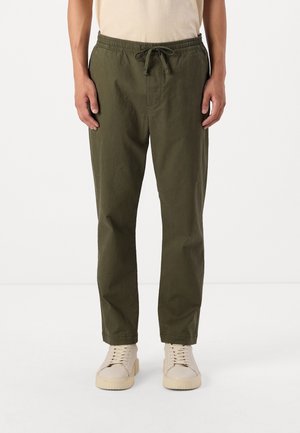 Mand iført olivengrønne snørebukser og beige sneakers, der står mod en ensfarvet hvid baggrund.