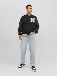 Marine varsityjack met witte strepen en een gouden 'N'-logo, gecombineerd met lichtblauwe jeans en zwart-witte sneakers. Glad textuur van de stof.