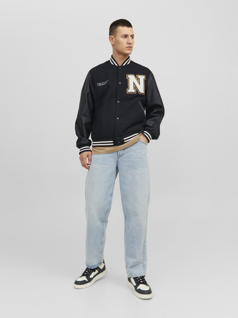 Chaqueta varsity navy con rayas blancas y logo dorado 'N', combinada con jeans azul claro y zapatillas en blanco y negro. Textura de tela suave.