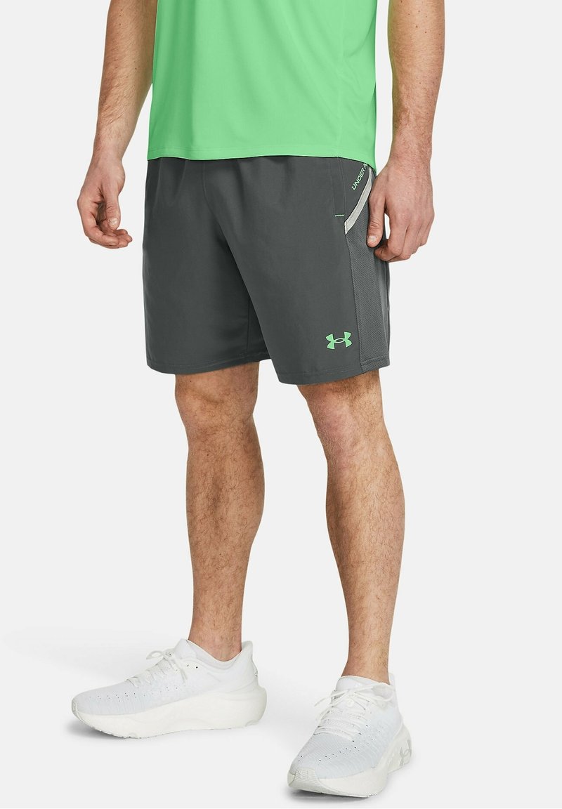 Šedé sportovní šortky s bočními síťovinovými panely, zelenými akcenty a logem Under Armour. V kombinaci s bílými sportovními botami.