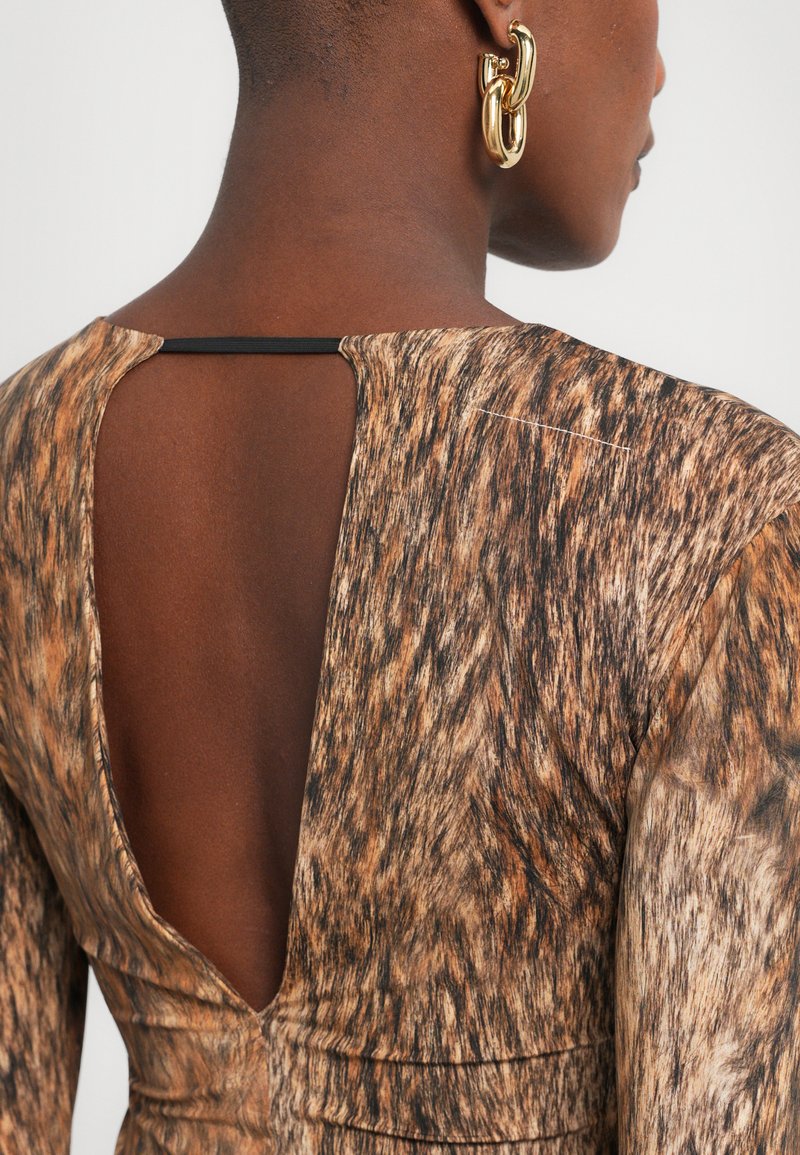 Närbild av en brun mönstrad klänning med djup V-rygg och strukturerad tyg. Ett guldörhänge är synligt i förgrunden.