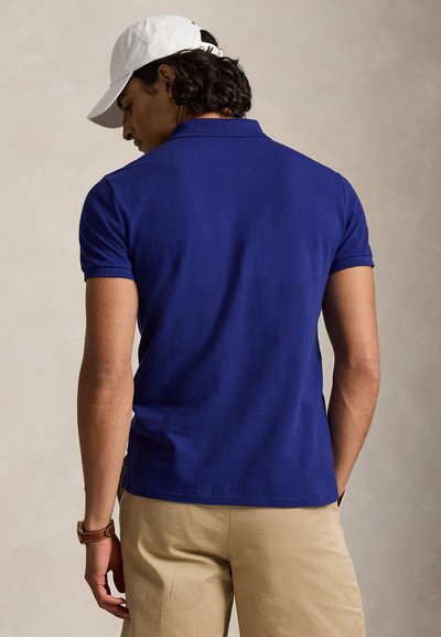 Mann, der ein blaues kurzärmeliges Poloshirt, beige Hose, weiße Kappe und braune Uhr trägt, von hinten vor einem einfarbigen Hintergrund gesehen.