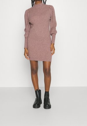 Femme portant une robe pull en maille côtelée mauve avec des manches longues bouffantes et des bottines noires épaisses, debout devant un fond uni.