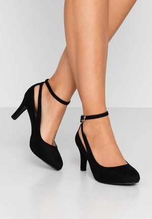 Des jambes portant des escarpins en suède noir avec des brides de cheville et une boucle en argent, se tenant sur un sol lisse de couleur claire.