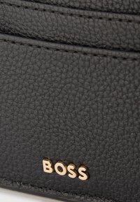 Surface en cuir noir texturé avec lettres métalliques dorées BOSS embossées près du bord cousu.