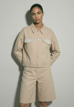 Kvinde med hænderne bag ryggen iført beige jakke med blonderlommer og matchende knælange shorts, stående mod en lys væg.
