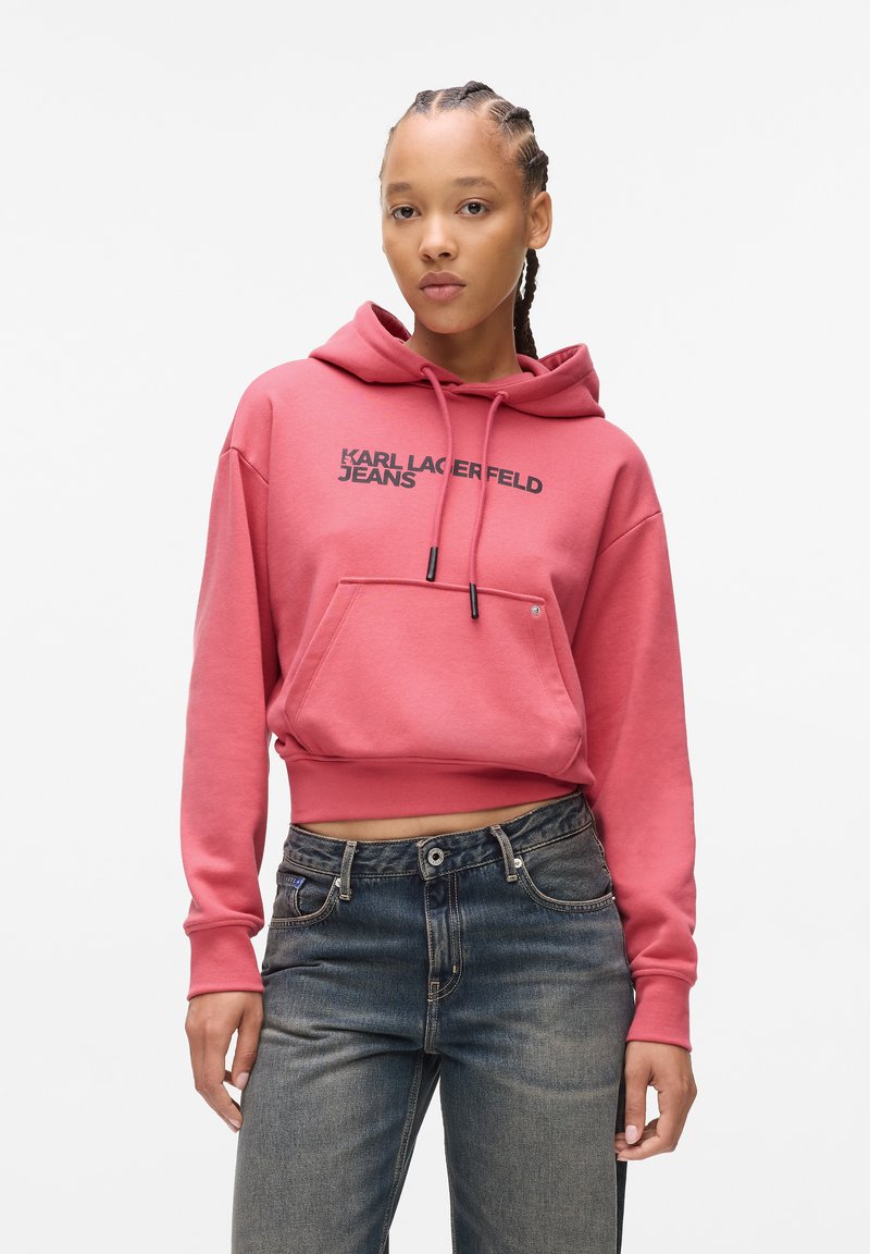 Rožinė trumpa apykaklė pagaminta iš minkštos medžiagos, su priekinėmis kišenėmis ir raišteliais. Ant jos yra juodas logotipo tekstas "KARL LAGERFELD JEANS."