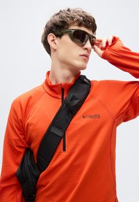 Πορτοκαλί πουκάμισο με φερμουάρ, μακρύ μανίκι με υφή· μαύρη τσάντα χιαστί· πράσινα γυαλιά ηλίου· μοντέλο που αγγίζει τα γυαλιά, ουδέτερο φόντο.