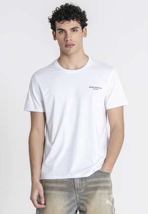 Joven con cabello rizado que lleva una camiseta blanca de Gianni Kavanagh Atelier y vaqueros grises descoloridos, de pie con una mano en el bolsillo.