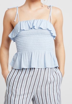 Hellblaues, gerafftes Oberteil mit Rüschenbesatz und Bindebändern, kombiniert mit hochtaillierten, gestreiften Hosen, Hände in den Taschen.